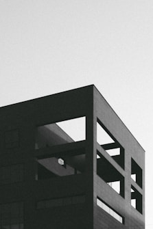 A modern, minimalistic building with a geometric design featuring sharp angles and clean lines. The facade is composed of square and rectangular shapes, and the image is rendered in black and white, lending it a stark and moody aesthetic.