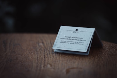 A close-up of a small informational card placed on a wooden surface. The card is slightly folded, displaying text in German. Water droplets are visible on the card, suggesting it is wet or has been exposed to rain. The background is dark, creating a contrast with the lighter card.