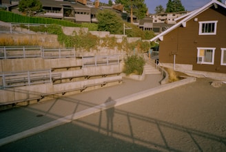 A terraced outdoor space with concrete steps and metal railings, featuring lush greenery along the upper concrete wall. In the background, residential buildings are partly visible behind trees. A brown building with white-trimmed windows is to the right. Shadows stretch across the foreground, indicating a low sun angle.