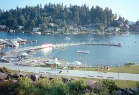 A scenic view of a lakeside area with a circular pier extending into the water. People are enjoying leisurely activities like swimming and sunbathing. Boats are docked at a marina to the left, and residential houses are nestled among dense forests in the background. A grassy area with paths and benches is in the foreground, offering spots for relaxation.