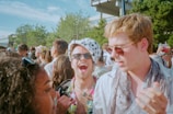A lively outdoor scene with a diverse group of people gathered together, suggesting a social event or festival. Many are wearing sunglasses and light clothing, indicating a sunny day. One person stands out with a spotted hat and is laughing enthusiastically.