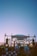 A large outdoor music festival during sunset with a stage set up featuring lights and decorations. A crowd of people in hats and vibrant clothing is gathered in front of the stage, enjoying the performance. The sky above is clear with a hint of an airplane in the distance.