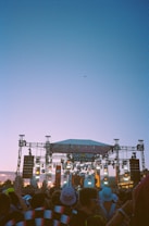 A large outdoor music festival during sunset with a stage set up featuring lights and decorations. A crowd of people in hats and vibrant clothing is gathered in front of the stage, enjoying the performance. The sky above is clear with a hint of an airplane in the distance.
