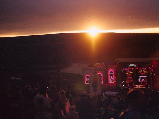 A sunset scene over a large outdoor music concert. The sun is setting behind a dark horizon, casting a warm, glowing light in the sky. Below the horizon, there is a stage illuminated with colorful lights and screens, while a crowd of people is gathered in the foreground, facing the stage.