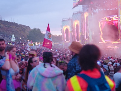 A large crowd of people gathered at an outdoor festival or concert. The scene is vibrant with many attendees in colorful clothing and accessories. Flags are waving above the crowd, and large illuminated stages can be seen in the background, with smoke or mist creating an atmospheric effect.