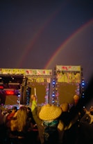 A nighttime concert scene with a crowd enjoying music under double rainbows. The stage has vibrant lights and decorations.