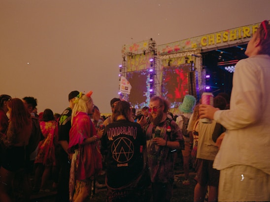 People are gathered at an outdoor music festival during the evening. They are dressed in colorful and eclectic clothing, with some wearing hats and holding drinks. A large stage with vibrant visual displays is visible in the background, adorned with bright lights and the name 'CHESHIRE' at the top.