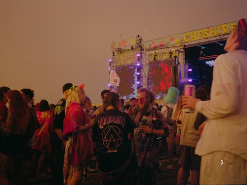 People are gathered at an outdoor music festival during the evening. They are dressed in colorful and eclectic clothing, with some wearing hats and holding drinks. A large stage with vibrant visual displays is visible in the background, adorned with bright lights and the name 'CHESHIRE' at the top.