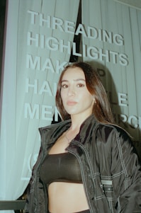 A person with long dark hair is standing in front of a sign with words like 'THREADING', 'HIGHLIGHTS', and 'MAKEUP'. The person is wearing a dark jacket and a black top. The image has an urban setting with vertical blinds visible in the background.