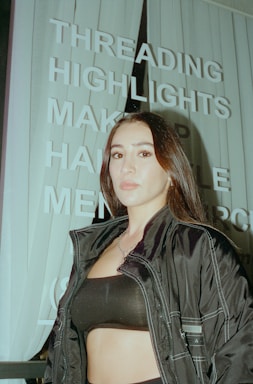 A person with long dark hair is standing in front of a sign with words like 'THREADING', 'HIGHLIGHTS', and 'MAKEUP'. The person is wearing a dark jacket and a black top. The image has an urban setting with vertical blinds visible in the background.