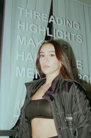 A person with long dark hair is standing in front of a sign with words like 'THREADING', 'HIGHLIGHTS', and 'MAKEUP'. The person is wearing a dark jacket and a black top. The image has an urban setting with vertical blinds visible in the background.