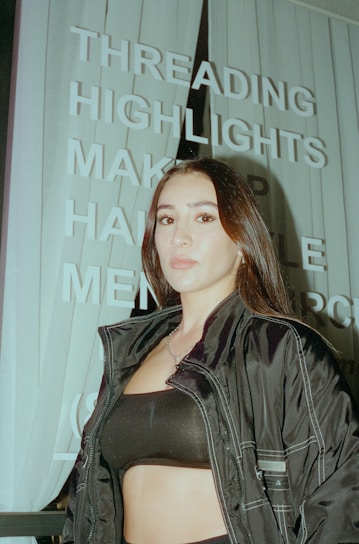 A person with long dark hair is standing in front of a sign with words like 'THREADING', 'HIGHLIGHTS', and 'MAKEUP'. The person is wearing a dark jacket and a black top. The image has an urban setting with vertical blinds visible in the background.