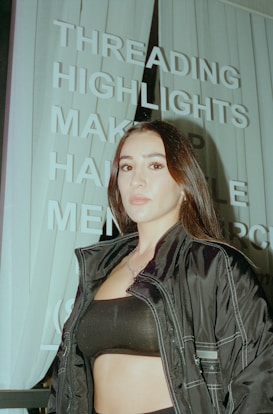 A person with long dark hair is standing in front of a sign with words like 'THREADING', 'HIGHLIGHTS', and 'MAKEUP'. The person is wearing a dark jacket and a black top. The image has an urban setting with vertical blinds visible in the background.