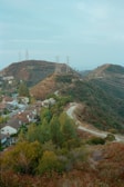 Rolling hills with a winding path and scattered green vegetation are seen with power lines stretching across the ridges. At the foot of the hills, a small residential area with closely packed houses featuring red-tiled roofs lies nestled among trees.
