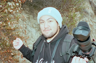 A person wearing a light blue knit beanie and a black hoodie, carrying a camera with a large lens mounted on a handheld stabilizer. They stand in front of a rocky background with small flowering plants. The person appears to be outdoors, possibly engaging in photography or videography.