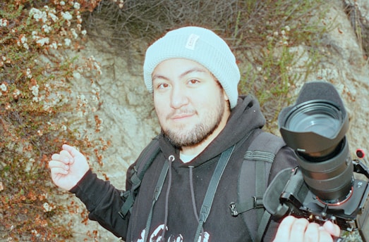 A person wearing a light blue knit beanie and a black hoodie, carrying a camera with a large lens mounted on a handheld stabilizer. They stand in front of a rocky background with small flowering plants. The person appears to be outdoors, possibly engaging in photography or videography.