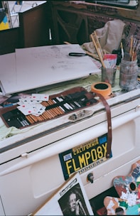 A cluttered artistic workspace with various art supplies including pencils, brushes, and containers. An unfinished sketch is on paper, surrounded by playing cards. An old car license plate from California is visible, along with a newspaper clipping of a person performing.