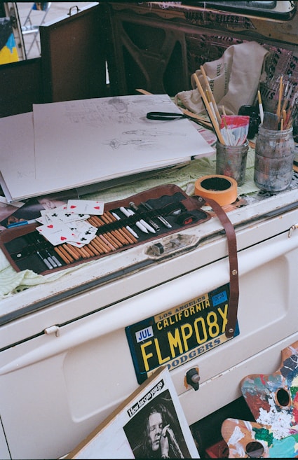 A cluttered artistic workspace with various art supplies including pencils, brushes, and containers. An unfinished sketch is on paper, surrounded by playing cards. An old car license plate from California is visible, along with a newspaper clipping of a person performing.