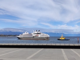 A large white cruise ship is seen on a calm body of water with a smaller yellow tugboat nearby. The distant background displays a range of blue mountains under a partly cloudy sky. The foreground features a concrete promenade leading towards the water.