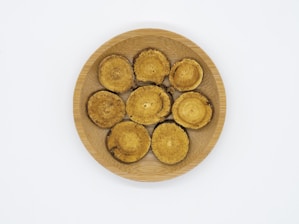 Seven circular, beige and brown objects that appear to be dried mushroom slices are arranged on a round, wooden plate. The background is plain white.