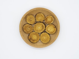 Seven circular, beige and brown objects that appear to be dried mushroom slices are arranged on a round, wooden plate. The background is plain white.