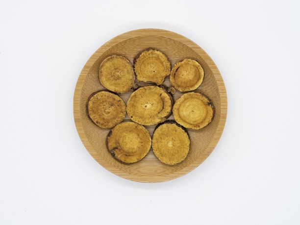 Seven circular, beige and brown objects that appear to be dried mushroom slices are arranged on a round, wooden plate. The background is plain white.