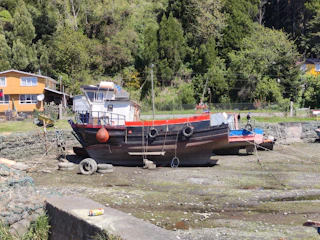 A boat is resting on a dry dock with a background of lush green trees. The boat has a mainly black hull with red and blue accents, and there are tires hanging on the sides. A house with yellow siding is visible in the background, and a discarded can is on a cement surface in the foreground. The scene is set in a rural, natural environment.