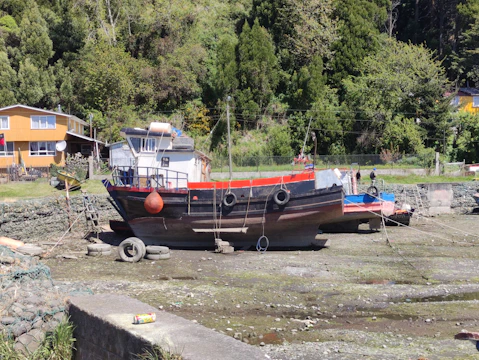A boat is resting on a dry dock with a background of lush green trees. The boat has a mainly black hull with red and blue accents, and there are tires hanging on the sides. A house with yellow siding is visible in the background, and a discarded can is on a cement surface in the foreground. The scene is set in a rural, natural environment.