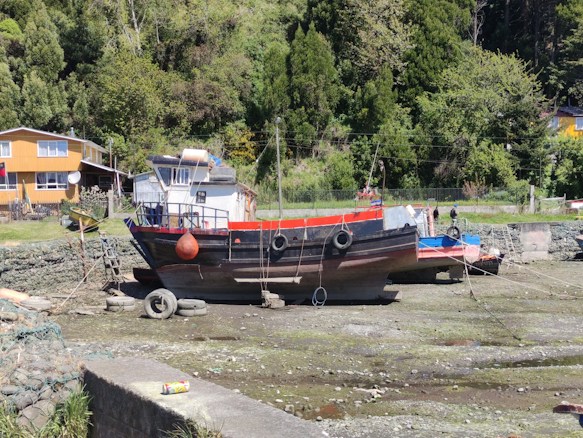 A boat is resting on a dry dock with a background of lush green trees. The boat has a mainly black hull with red and blue accents, and there are tires hanging on the sides. A house with yellow siding is visible in the background, and a discarded can is on a cement surface in the foreground. The scene is set in a rural, natural environment.