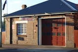 A brick building with a large garage door and a sign reading 'Fire + Rescue'. The structure has a dark sloped roof, a window with white panes, and an Australian flag visible in the background. The sunlight casts shadows on the brick wall.