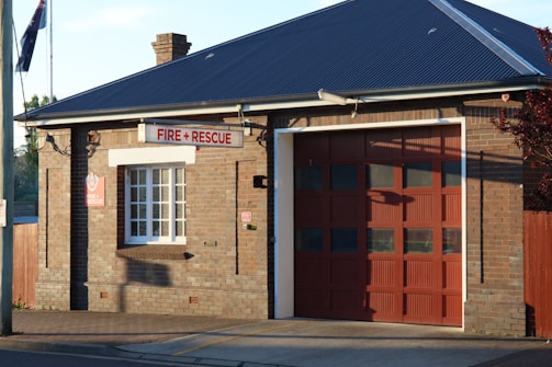A brick building with a large garage door and a sign reading 'Fire + Rescue'. The structure has a dark sloped roof, a window with white panes, and an Australian flag visible in the background. The sunlight casts shadows on the brick wall.