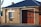 A brick building with a large garage door and a sign reading 'Fire + Rescue'. The structure has a dark sloped roof, a window with white panes, and an Australian flag visible in the background. The sunlight casts shadows on the brick wall.