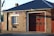 A brick building with a large garage door and a sign reading 'Fire + Rescue'. The structure has a dark sloped roof, a window with white panes, and an Australian flag visible in the background. The sunlight casts shadows on the brick wall.