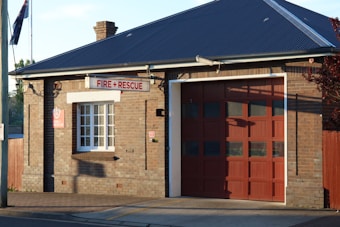 A brick building with a large garage door and a sign reading 'Fire + Rescue'. The structure has a dark sloped roof, a window with white panes, and an Australian flag visible in the background. The sunlight casts shadows on the brick wall.