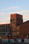 A tall brick building with several windows, some rectangular and others covered with metal panels. In front of the building, there are multiple dumpsters labeled JR Richards. To the side, scaffolding is present, suggesting construction or renovation work is underway. The sky is blue with a few scattered clouds.