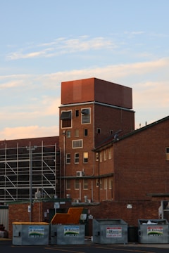 A tall brick building with several windows, some rectangular and others covered with metal panels. In front of the building, there are multiple dumpsters labeled JR Richards. To the side, scaffolding is present, suggesting construction or renovation work is underway. The sky is blue with a few scattered clouds.