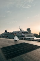 A cruise ship is docked beside a wooden boardwalk, where a person in a white dress is holding a bouquet and appears to be posing. The sky is clear with scattered clouds, and buildings can be seen in the background, contributing to a peaceful atmosphere.
