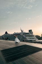 A cruise ship is docked beside a wooden boardwalk, where a person in a white dress is holding a bouquet and appears to be posing. The sky is clear with scattered clouds, and buildings can be seen in the background, contributing to a peaceful atmosphere.