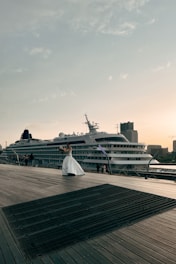 A cruise ship is docked beside a wooden boardwalk, where a person in a white dress is holding a bouquet and appears to be posing. The sky is clear with scattered clouds, and buildings can be seen in the background, contributing to a peaceful atmosphere.
