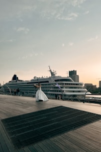 A cruise ship is docked beside a wooden boardwalk, where a person in a white dress is holding a bouquet and appears to be posing. The sky is clear with scattered clouds, and buildings can be seen in the background, contributing to a peaceful atmosphere.