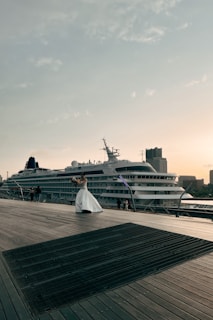 A cruise ship is docked beside a wooden boardwalk, where a person in a white dress is holding a bouquet and appears to be posing. The sky is clear with scattered clouds, and buildings can be seen in the background, contributing to a peaceful atmosphere.