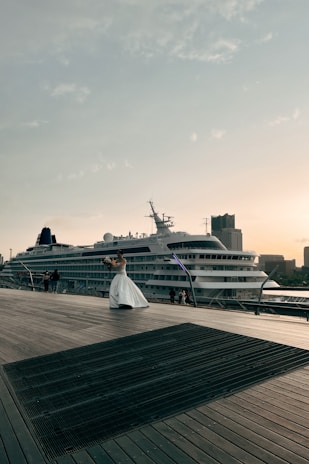 A cruise ship is docked beside a wooden boardwalk, where a person in a white dress is holding a bouquet and appears to be posing. The sky is clear with scattered clouds, and buildings can be seen in the background, contributing to a peaceful atmosphere.