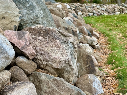 A pile of various sized rocks forms a rustic stone wall alongside a grassy area. The stones are predominantly grey and brown, with some showing pinkish hues, and are surrounded by scattered dried leaves. The grass is lush and vibrant, indicating a natural, outdoor setting.
