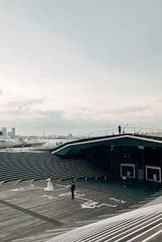A couple, with the woman wearing a flowing white gown, stands on an expansive wooden deck with architectural steps and a modern building in the background. The wide open sky and city skyline are visible in the distance under a cloudy sky. A person is also seen on the roof of the building taking pictures.