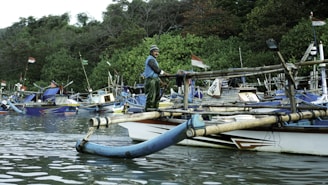 A person stands on a traditional wooden boat in a calm body of water, surrounded by other similar boats. The boats are equipped with bamboo outriggers and are tied up near a dense, green forested shoreline. Indonesian flags are visible on the boats.