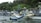 A person stands on a traditional wooden boat in a calm body of water, surrounded by other similar boats. The boats are equipped with bamboo outriggers and are tied up near a dense, green forested shoreline. Indonesian flags are visible on the boats.