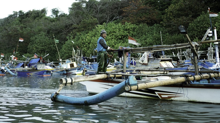 A person stands on a traditional wooden boat in a calm body of water, surrounded by other similar boats. The boats are equipped with bamboo outriggers and are tied up near a dense, green forested shoreline. Indonesian flags are visible on the boats.