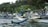 A person stands on a traditional wooden boat in a calm body of water, surrounded by other similar boats. The boats are equipped with bamboo outriggers and are tied up near a dense, green forested shoreline. Indonesian flags are visible on the boats.