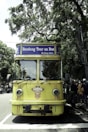 A vintage-style yellow bus with 'Bandung Tour on Bus' written on the front, parked on a tree-lined street. The bus has round headlights and a distinctive front design. Several people are standing nearby, some wearing masks. The surrounding environment is lush with green trees, and there's a streetlamp with a globe light.