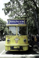A vintage-style yellow bus with 'Bandung Tour on Bus' written on the front, parked on a tree-lined street. The bus has round headlights and a distinctive front design. Several people are standing nearby, some wearing masks. The surrounding environment is lush with green trees, and there's a streetlamp with a globe light.